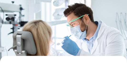 Ein Zahnarzt, der eine Maske, Handschuhe und eine Schutzbrille trägt, untersucht einen Patienten, der auf einem Zahnarztstuhl in einer hellen Zahnklinik sitzt.