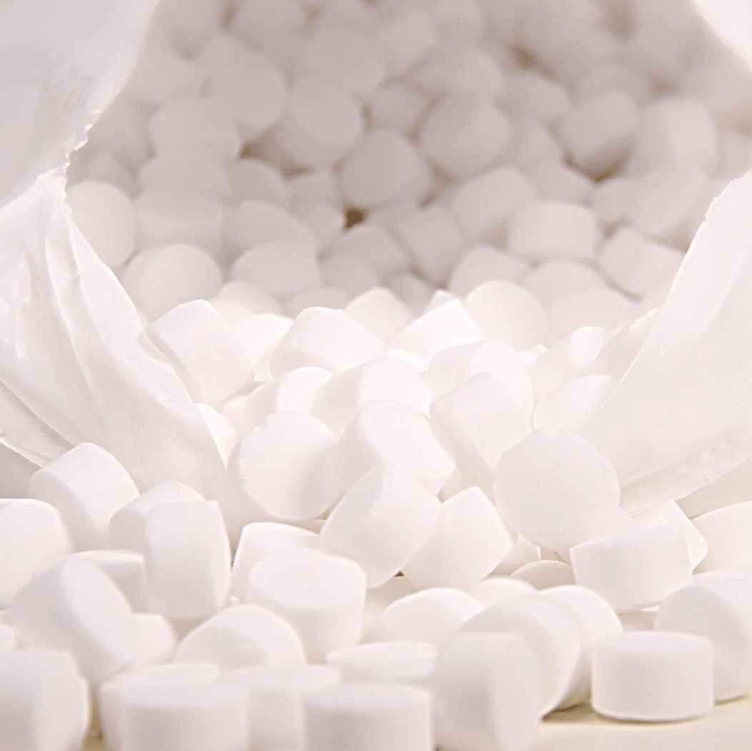 Et nærbillede viser en hvid plastpose med påskriften »K+S Minerals and Agriculture GmbH«, der indeholder Regenit-kogesaltstabletter i henhold til EN 973, type | sæk (25 kg), som falder ned på en overflade. De små, runde tabletter har en ensartet form og størrelse og er ideelle til brug i vandblødgøringsanlæg.