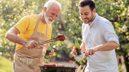 Zwei Männer grillen gemeinsam im Garten, halten Grillzange und Getränke in der Hand, lachen vor unscharfem Hintergrund