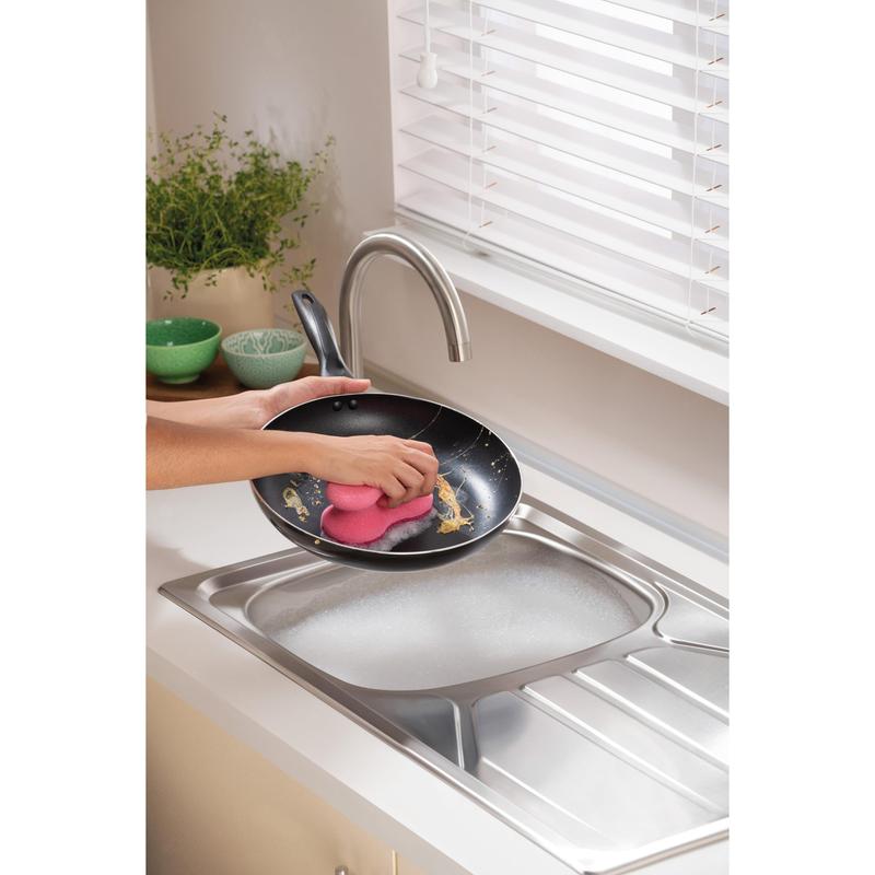 En person rengør en stegepande under rindende vand i et moderne køkken med en 3M Deutschland GmbH Scotch-Brite® Delicate Color Comfort ikke-ridsende rengøringssvamp (2 stk.) - ideel til sarte overflader. Grønne skåle og en plante står på bordpladen.