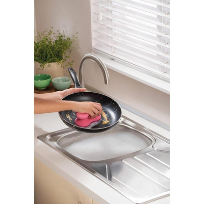 En person rengør en stegepande under rindende vand i et moderne køkken med en 3M Deutschland GmbH Scotch-Brite® Delicate Color Comfort ikke-ridsende rengøringssvamp (2 stk.) - ideel til sarte overflader. Grønne skåle og en plante står på bordpladen.