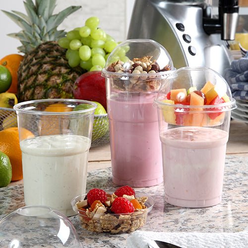 Tre smoothies i PAPSTAR GmbH kolddrikbæger PLA "pure" glasklar 0,5 l står på en køkkenbordplade. To har frugter og müsli under kuppellåg. I baggrunden ses friske frugter og en blender. Pakken indeholder 60 stykker.