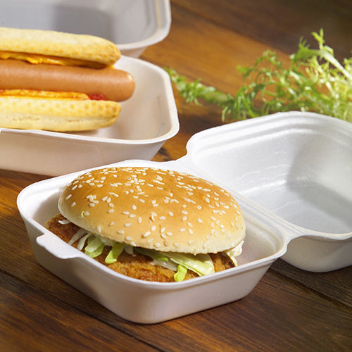 Ein Hamburger in einem Sesambrötchen mit Salat und Patty, serviert in einer Starpak 100 Hamburger-Box mit Klappdeckel, XPS, 7,5 x 12 x 12 cm weiß – ideal fürs Catering, mit Hotdogs in der Nähe und Blattgemüse auf einer Holzoberfläche.
