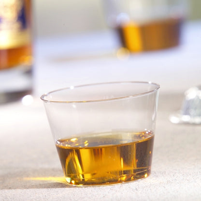 En Starpak 15 drikkebæger, PS 0,1 l krystalklar, indeholder en lille mængde ravfarvet væske på en lys overflade, mens en anden kop og en flaske er slørede i baggrunden.