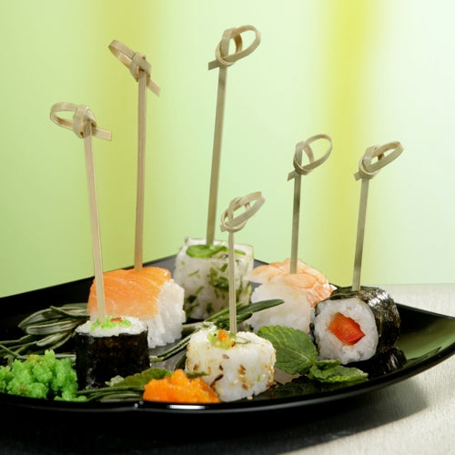 Auf einem schwarzen Teller liegen verschiedene Sushi-Rollen, aufgespießt mit jeweils einem PAPSTAR 250 Fingerfood - Picker „pure“ der PAPSTAR GmbH, arrangiert mit grünen Garnituren vor einem unscharfen gelbgrünen Hintergrund.