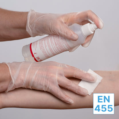 Eine Person in ARNOMED VINYL PF Tastsensible Einmalhandschuhe von ARNOWA GmbH benutzt eine Sprühflasche, um ein Wattepad zu befeuchten und den Unterarm einer anderen Person zu reinigen. In der unteren rechten Ecke erscheint das Etikett "EN 455".