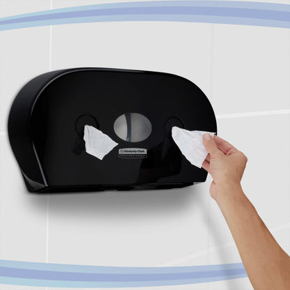 En person trækker toiletpapir ud af en sort Kimberly-Clark Professional™ toiletpapirdispenser - centraludtag (Jumbo) fra Kimberly-Clark GmbH, som er monteret på en flisevæg med blå buede striber.