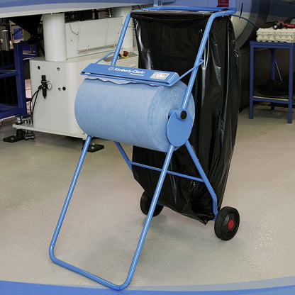 En Kimberly-Clark Professional™ Mobilstander til aftørringsruller (Storrulle/Blå) med en rulle blå industripapirhåndklæder og en stor sort affaldspose står på hjul på et gråt lagergulv ved en arbejdsplads.