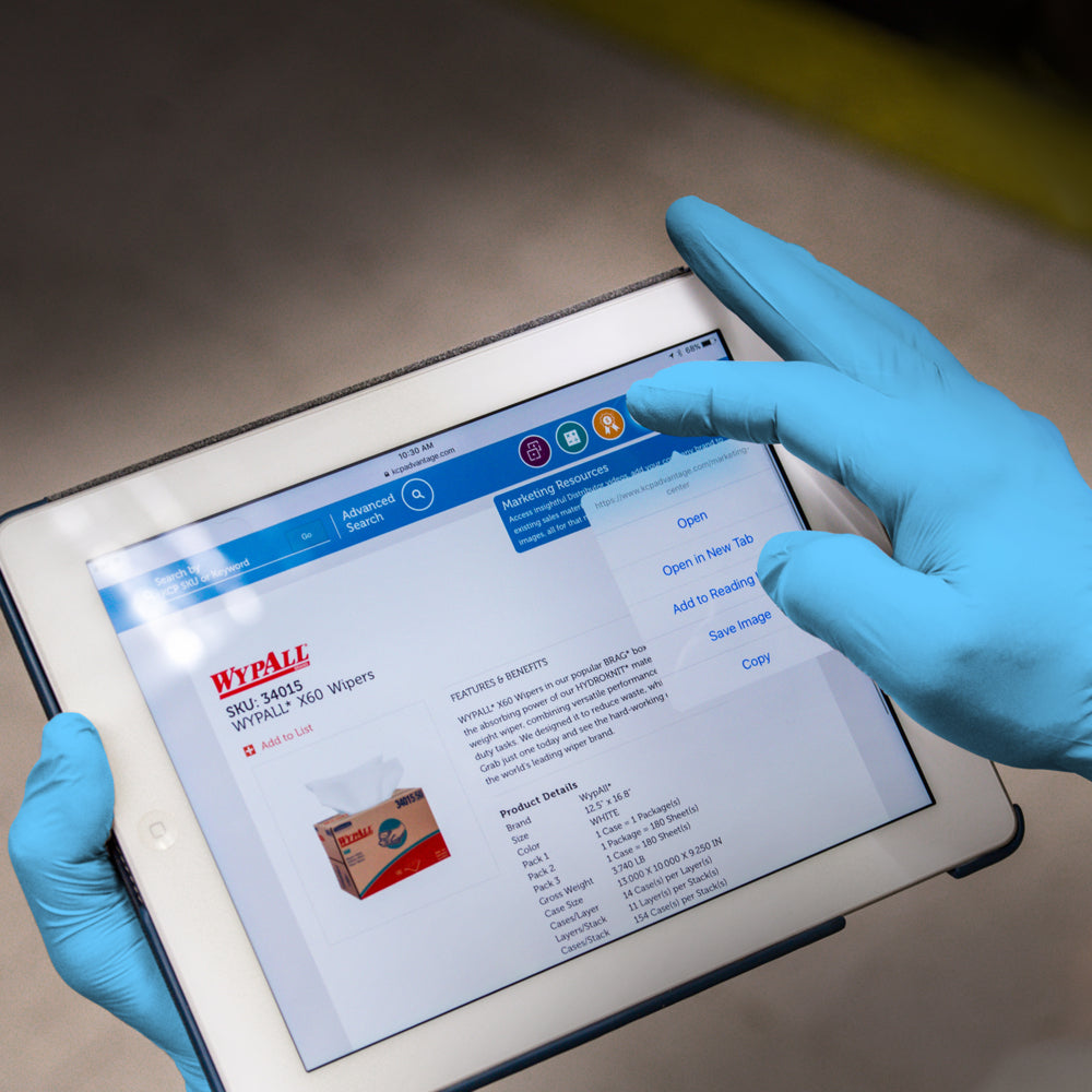 Eine Person, die KleenGuard® G10 Comfort Plus Blaue Nitril-Handschuhe von Kimberly-Clark GmbH trägt, hält ein Tablet, das eine Produktseite mit Bild, Beschreibung und Kontextmenü zeigt; der Hintergrund ist unscharf.