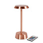 Den trådløse NOUR LED-lampe i roséguld fra Duni GmbH har en rund base og en flad top for en moderne stil. Ved siden af lampen ligger en fjernbetjening med farverige knapper.