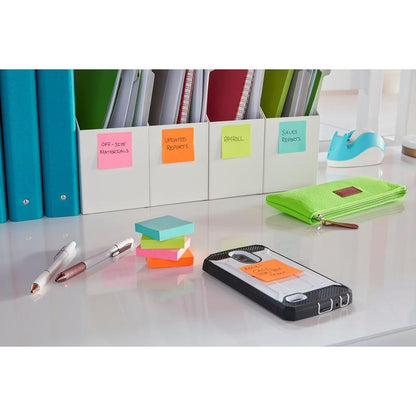 På det ryddelige skrivebord ligger mærkede mapper, en grøn mappe, to penne og en smartphone med en Post-it® Super Sticky Note (76x127 mm, 3M Deutschland GmbH), hvorpå der står "AM Call Client Chet". Den godt oplyste overflade er hvid.