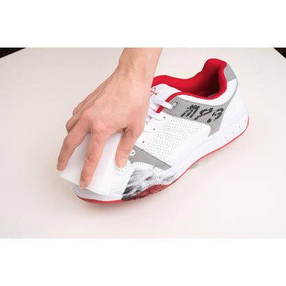 En hånd anvender Scotch-Brite® Magic Pad fra 3M Deutschland GmbH til at fjerne sorte ridsemærker fra siden af en hvid sneaker med røde og grå accenter på en hvid overflade.
