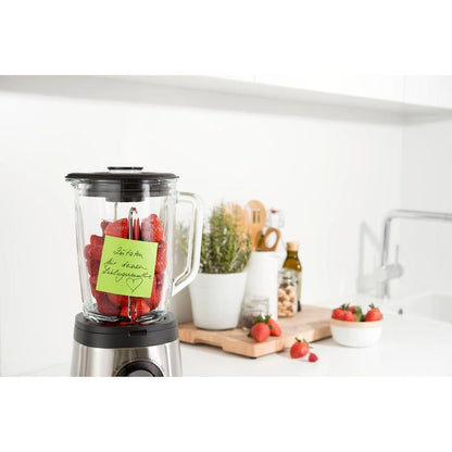 En blender med jordbær står på en hvid køkkenbordplade. En Post-it® Super Sticky Note (76 x 76 mm, 90 ark, 100% PEFC) fra 3M Deutschland GmbH er fastgjort til beholderen. I nærheden er redskaber, potteplanter, olivenolie og flere jordbær på et skærebræt.