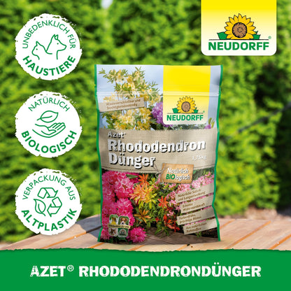 En pose Azet RhododendronGødning fra Neudorff Shop ligger på en træflade udendørs foran grønne hække. På emballagen af bioplast står der, at den er biologisk, kæledyrsvenlig og ideel til rhododendron og azalea.