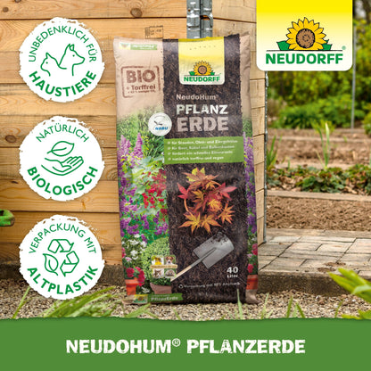 En pose NeudoHum Plantjord fra Neudorff Shop står oprejst i en have, perfekt til stauder og buske. Symbolerne viser, at den er biologisk, dyreforsøgfri, tørvefri og fremstillet af genanvendt plastik; Neudorff-logoet er placeret i øverste højre hjørne.