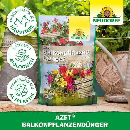 En pose Azet BalkonpflanzenGødning fra Neudorff Shop står på et bord mellem potteplanter og altankasser. Emballagen viser blomster, tekst og symboler for dyrebeskyttelse, biologiske ingredienser og emballage af bioplast.