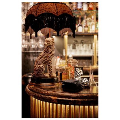 Eine schwach beleuchtete Bar mit einer Leopardenstatue auf der Theke unter einem Lampenschirm mit Fransen besticht durch modernes Design. Flaschen und Servietten umgeben einen schicken Kerzenhalter Twine 75 x Ø 75 mm Schwarz von Duni GmbH, neben einer Metalllaterne entsteht eine einladende Tischdekoration.
.