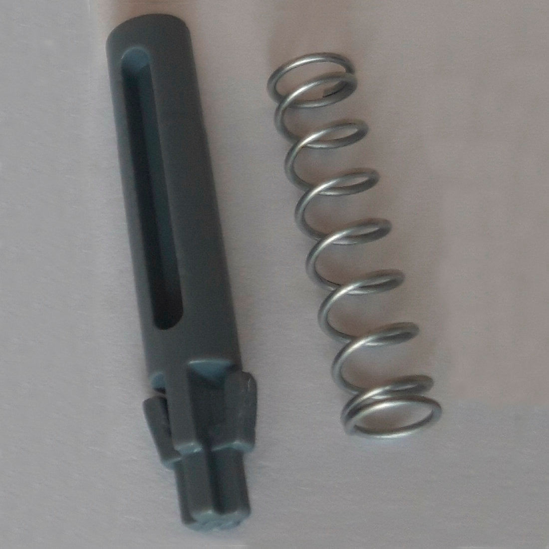 En metalfjeder og en mørkegrå cylindrisk reserve-dørstift med riller og takker, en del af DI Protect dørstift & fjeder (1 stk.) fra Diversey Deutschland GmbH & Co. OHG, er afbildet på en lys overflade og viser de robuste materialer til DI Protect Trolley.