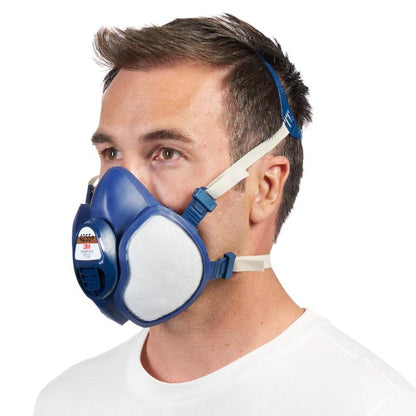En mand med kort brunt hår bærer en blå-hvid 3M™ åndedrætsmaske med integrerede filtre fra 3M Deutschland GmbH med justerbare stropper, afbildet foran en enkel hvid baggrund.