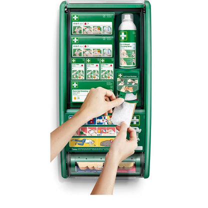 En person holder en forbinding foran en Cederroth førstehjælpsstation fra Orkla Wound Care AB, der er udstyret med forbindinger, brandskadegel, saltvandsopløsning og bandager. Illustrerede instruktioner kan ses på hylderne indeni.