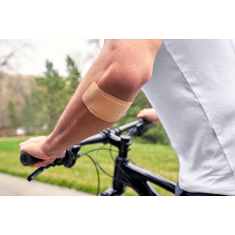 En person, der bærer en hvid skjorte og cykler udendørs, har et Nexcare™ Ultra Stretch MAXI Comfort Flexible plaster (50 mm x 101 mm) fra 3M Deutschland GmbH på den bare overarm; i baggrunden ses grønt græs og træer.