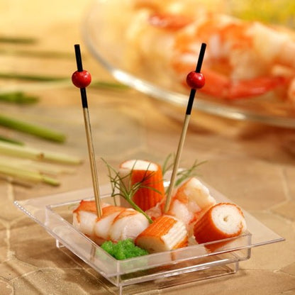 På en gennemsigtig, firkantet skål er der krabbeimitat, rejer, en grøn pynt og to PAPSTAR 250 fingerfood-pinde fra PAPSTAR GmbH med røde perler. I baggrunden kan man se slørede rejer på en tallerken.