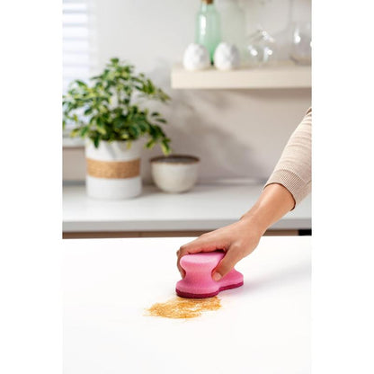 En hånd bruger en lyserød Scotch-Brite® Delicate Color Comfort ikke-ridsende rengøringssvamp (2 stk., 3M Deutschland GmbH) til at rengøre brunt pulver fra en hvid bordplade - perfekt til sarte overflader. Plante og skåle i baggrunden.