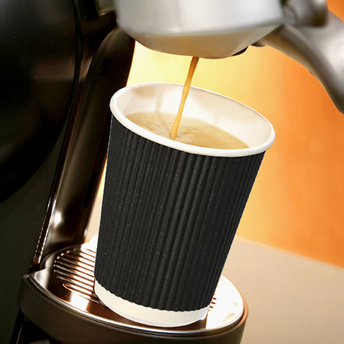 En Starpak 25 drikkekop, pap "To Go" Ripple Wall-kop står på en metaldrypbakke under udløbet af en kaffemaskine, mens varm kaffe løber ind i den, og skiller sig ud foran en blidt oplyst, varmtonet baggrund.