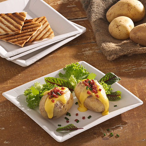 To bagte kartofler med gul ostesauce, baconstykker og purløg serveres på en PAPSTAR pap-tallerken firkantet 22,5 cm i "pure" design af PAPSTAR GmbH. Bladgrøntsager ledsager retten, i baggrunden står grillet brød og hele kartofler.