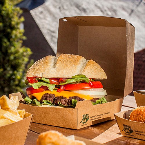 En cheeseburger med salat, tomat, løg og cheddarost ligger udenfor i en PAPSTAR GmbH Burgeræske af pap (9x18,5x18,5 cm, »100 % Fair«, 25 stk.). Pommes frites og andre tilbehør serveres separat ved siden af.