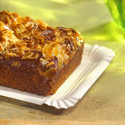 På den PAPSTAR pap-tallerken firkantet 13x20 cm, hvid, "pure" til fester (12 styk) fra PAPSTAR GmbH hviler en rektangulær kage med flager af mandler - en miljøvenlig, elegant serveringsmulighed på en lys overflade med grønt glas bagved.