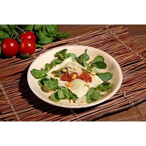 En tallerken ravioli med pesto, soltørret tomatsauce, pinjekerner og ost serveres på friskt grønt i en PAPSTAR palmebladsskål (17,5 cm), en miljøvenlig skål fra det 6-delte sæt fra PAPSTAR GmbH, serveret med tomater og bladgrøntsager i baggrunden.