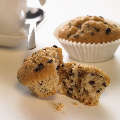 Im Vordergrund ist ein halbierter Chocolate Chip Muffin zu sehen, über den Krümel verstreut sind. Im Hintergrund liegt ein weiterer Muffin in einem PAPSTAR 1000 Backförmchen rund der PAPSTAR GmbH, neben einer weißen Tasse mit Untertasse und Löffel auf weißem Untergrund.