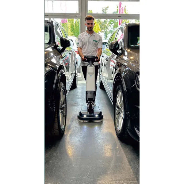 Eine Person im weißen Hemd setzt die Arcora Tiger 40 Bodenreinigungsmaschine von Arcora International GmbH zwischen zwei schwarzen Autos in einem hellen Innenraum mit großen Fenstern ein und sorgt für eine professionelle Reinigung.