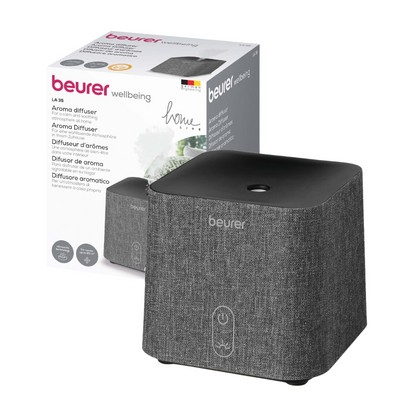 Billede af en Beurer LA 35 aroma-diffusor foran sin emballageæske. Diffusoren fra Beurer GmbH er grå med en stofstruktur og har en enkelt betjeningsknap på forsiden. Den bruger ultralydsforstøvning til en effektiv forstøvning, og på æsken er den samme diffusor, produktnavn og detaljer afbildet på flere sprog.