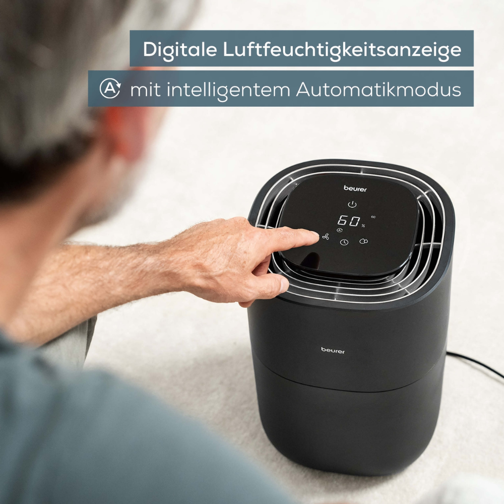 Man ser en person, der interagerer med en Beurer LB 200 luftfugter fra Beurer GmbH. Enhedens digitale display viser en luftfugtighed på 50 %. Den ledsagende tekst på tysk lyder „Digital luftfugtighedsvisning med intelligent automatisk tilstand“.