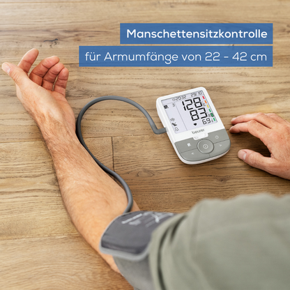 Ein Anwender arbeitet mit dem Beurer GmbH Auslaufartikel: Oberarm-Blutdruckmessgerät BM 53 auf einer hellen Holzoberfläche. Das Display zeigt "Manschettensitzkontrolle für Armumfänge von 22-42 cm" und weist auf Arrhythmieerkennung und Schlaganfallprävention hin.