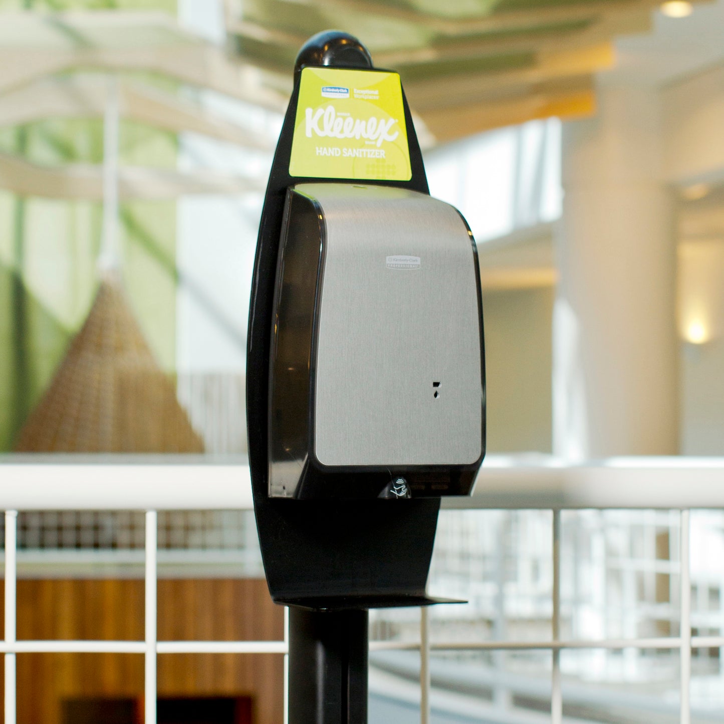 En Kimberly-Clark GmbH Elektroniske Vaskelotionsstander (Sort, 1 dispenser) er placeret indendørs nær et hvidt metalgelænder til hygiejnisk håndvask, med slørede arkitektoniske detaljer i baggrunden.