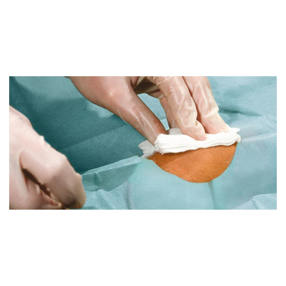 Et nærbillede af behandskede hænder, der renser et lille hudområde med en hvid klud, omgivet af blågrønne Hartmann Foliodrape® Protect hulklude, selvklæbende, steril, individuelt pakket, fra Paul Hartmann AG.
