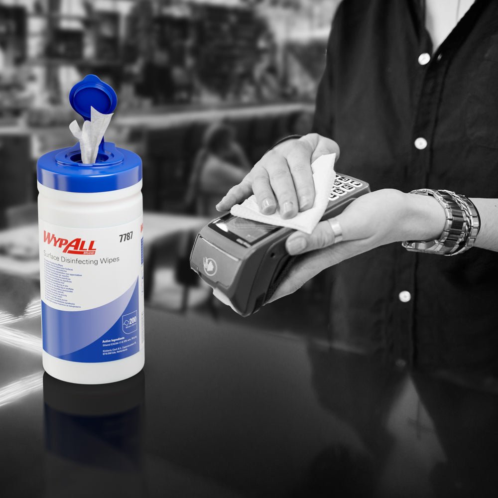 Nogen bruger en WypAll® overfladedesinfektionsserviet (Kimberly-Clark GmbH) fra en åben dispenserbakke til desinfektion af en kortterminal. Den hvide dispenser spand (200 servietter) står på disken, baggrunden er sløret.
