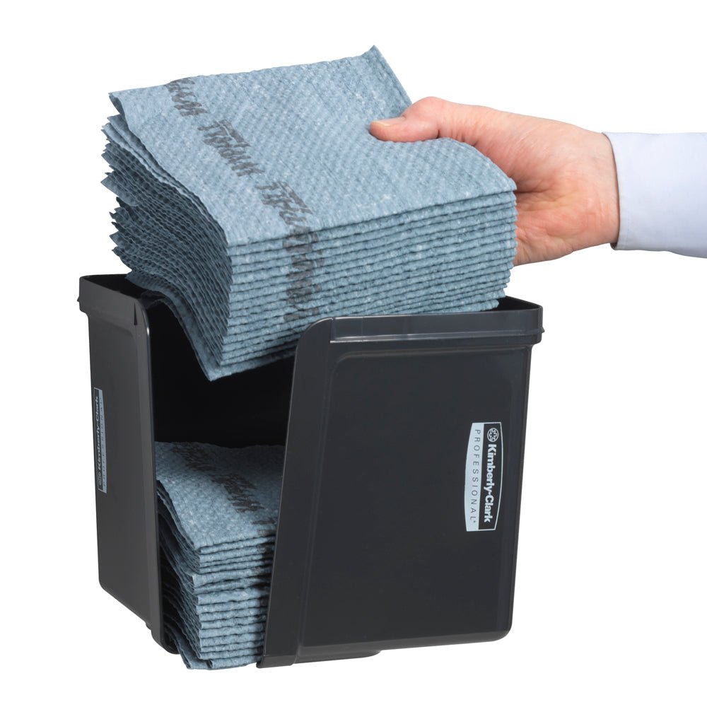 En hånd lægger kvartfoldede klude i den grå Kimberly-Clark Professional™ kluddispenser fra Kimberly-Clark GmbH - for hygiejnisk klududtagning. De pænt foldede klude og den forreste etiket er synlige.