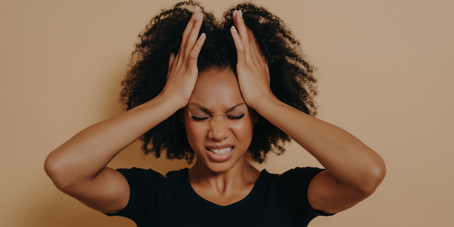 Eine Frau mit lockigem Haar und schwarzem Hemd presst ihre Hände an die Seiten ihres Kopfes und schließt fest die Augen, als wäre sie frustriert oder würde sich unwohl fühlen. Sie steht vor einem schlichten beigen Hintergrund.