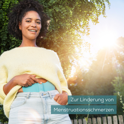 En person i en gul sweater og jeans står udenfor og holder en Beurer EM 55 Menstrual Relax+ gel-pude fra Beurer GmbH i hånden. I baggrunden ses grønt, gennem hvilket sollyset falder. Teksten lyder 'Til lindring af menstruationssmerter'.