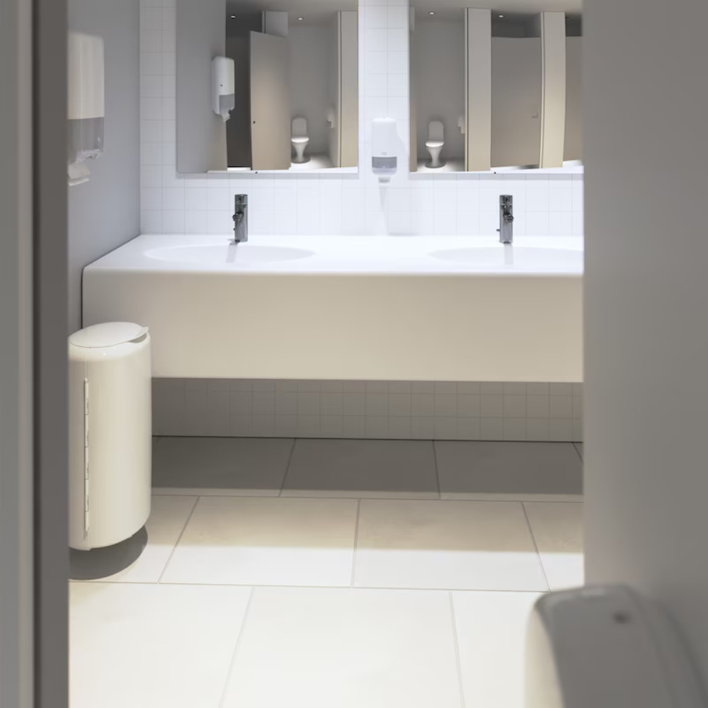 Die öffentliche Toilette präsentiert sich in modernem Elevation-Design mit weiß gefliesten Wänden und Böden. Eine lange weiße Arbeitsplatte beherbergt zwei Waschbecken unter einem großen Spiegel sowie einen Tork 553000 Spender für Zickzack-Handtücher Elevation H3 von TORK – Essity Professional Hygiene Germany GmbH, der eine effiziente Einzelblattausgabe für Papierhandtücher bietet. Ein praktisch positionierter Mülleimer befindet sich in der Nähe des Eingangs.