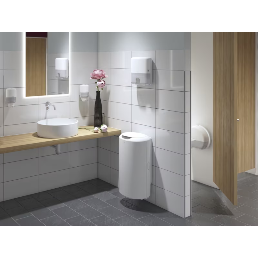 Eine moderne öffentliche Toilette mit weiß gefliesten Wänden und dunkelgrauem Boden ist ein Highlight des Elevation-Designs. Sie verfügt über eine Holzablage mit rundem Waschbecken, großem Spiegel, Tork 555000 Spender für Mini-Jumbo-Toilettenpapier Elevation T2 von Essity Professional Hygiene Germany GmbH, sowie einen Seifenspender, Handtuchspender, Blumenvase, Abfalleimer und eine teilweise geöffnete Kabinentür.