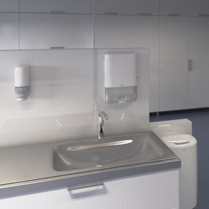 Vises er en moderne håndvask på et offentligt toilet med automatisk vandhane. Over håndvasken er en Tork Xpress® 552000 dispenser til multifold håndklæder H2 fra Essity Professional Hygiene Germany GmbH og en sæbedispenser monteret på en flisevæg. I baggrunden ses en række hvide skabe.