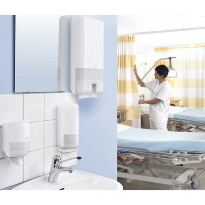 I et hospitalsrum står en læge i hvid uniform ved siden af en seng. I forgrunden er en håndvask udstyret med en Tork Xpress® 552000 dispenser til multifold håndklæder H2 fra TORK – Essity Professional Hygiene Germany GmbH. Rummet er udstyret med medicinsk udstyr og gule stribede gardiner.