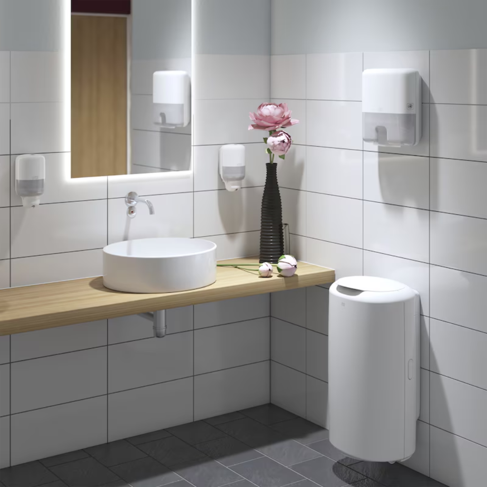 Ein modernes Badezimmer mit einem runden weißen Waschbecken auf einer Holzplatte, aufgewertet durch einen Wandspiegel mit Beleuchtung. Der Tork Xpress® 552100 Mini Spender für Multifold Handtücher Elevation H2 von TORK – Essity Professional Hygiene Germany GmbH verleiht den weiß gefliesten Wänden Eleganz, zusammen mit einer schwarzen Vase voller rosa Blumen. Auf dem grau gefliesten Boden steht ein weißer Abfalleimer.