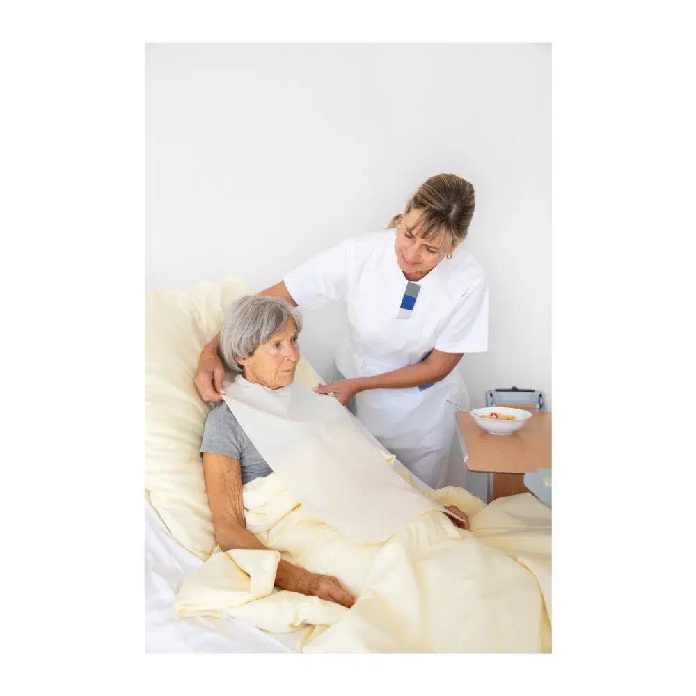 En sygeplejerske retter tæppet for en ældre kvinde, der sidder oprejst i en hospitalsseng. Begge ser opmærksomme og koncentrerede ud. En bakke med en skål og Vala® Fit Tape engangsbeklædningsbeskyttelse er synlig.