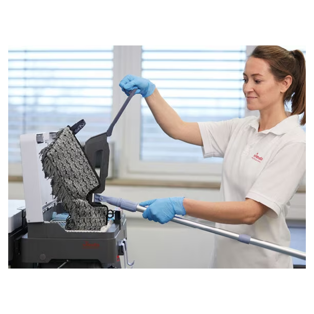 Eine Person mit blauen Handschuhen und weißem Hemd reinigt mit dem Vileda Professional Swep Duo r-MicroCombi Mop - 50 cm von Vileda GmbH eine strukturierte schwarze Oberfläche. Die Szene scheint in einem Innenbereich zu sein, mit einem großen Fenster im Hintergrund, durch das natürliches Licht in den Raum fällt.
